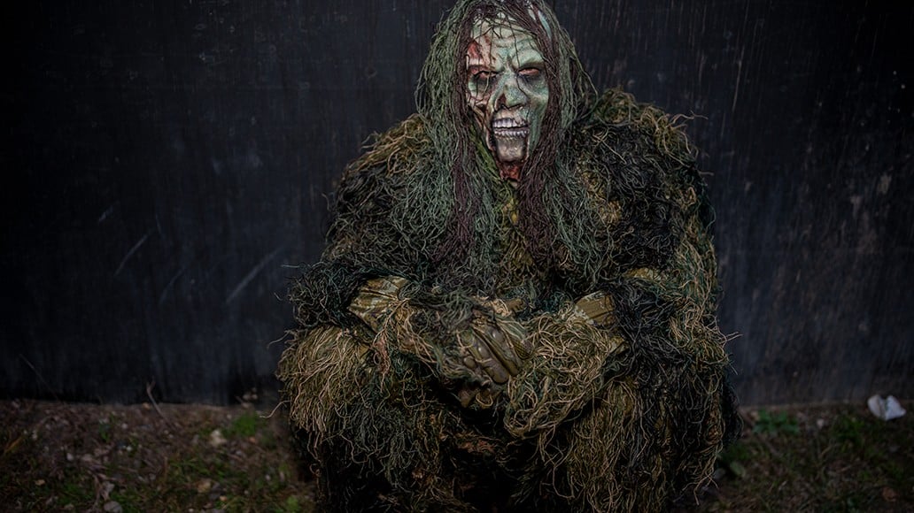 A performer in a moss-covered costume and a pale mask crouches on the ground in the dark