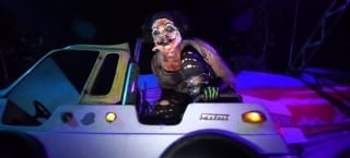 A costumed performer in pale face makeup sits on top of a bumper car under purple and blue light