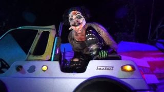 A costumed performer in pale face makeup sits on top of a bumper car under purple and blue light