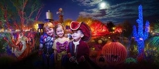 Three children in costumes, a superhero, a princess and a pirate, smile at night among glowing trees, a blue-lit cactus and a lit jack-o'-lantern at the Springs Preserve