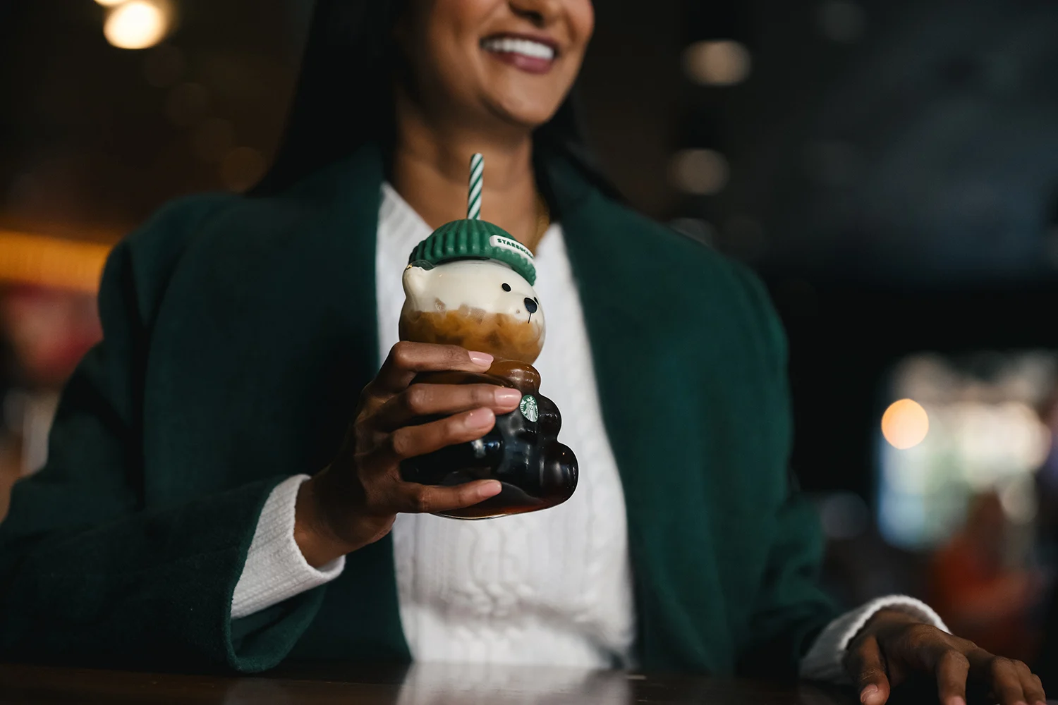 A customer holds the original Bearista Glass Cold Cup, a bear-shaped glass cup with a green hat, in a coffeehouse
