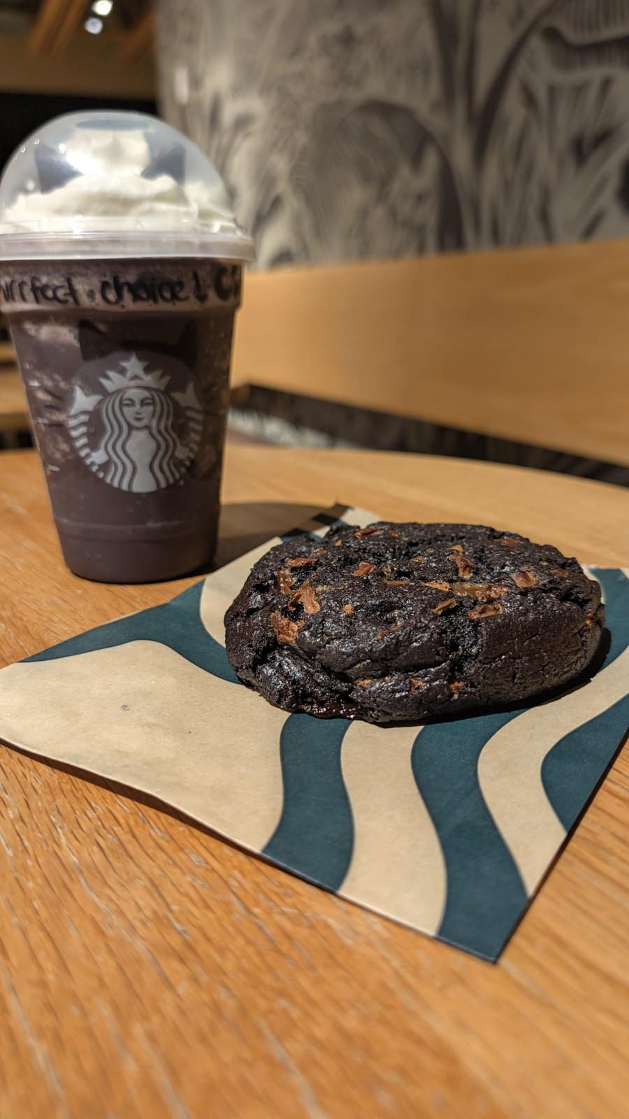 A Starbucks Black Cat Frappuccino beside a large black cocoa cookie with caramel pieces on a Starbucks napkin