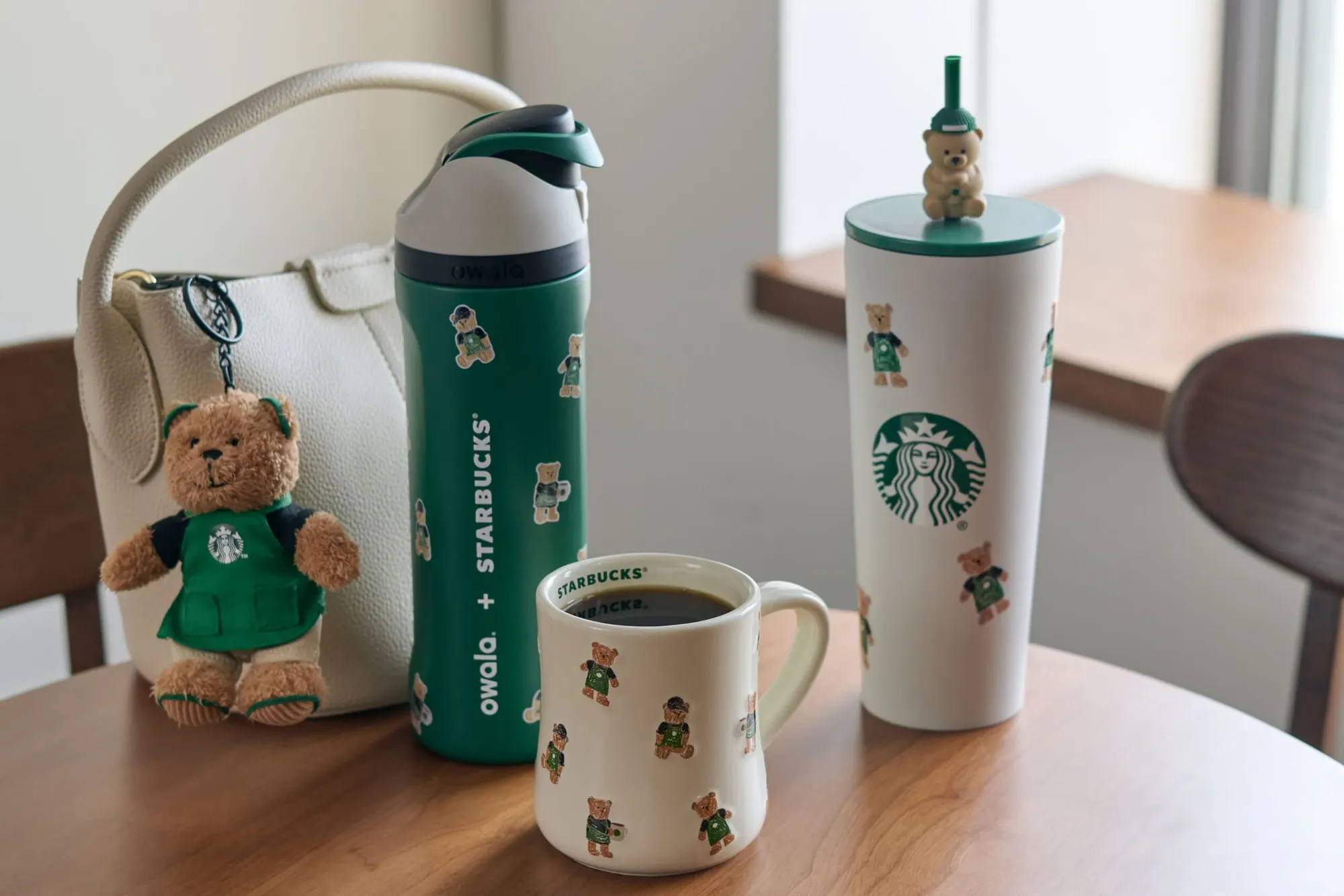Holiday Bearista pieces on a cafe table: a plush bear keychain on a tote, a green water bottle, a bear-print mug and a tumbler with a bear on the straw