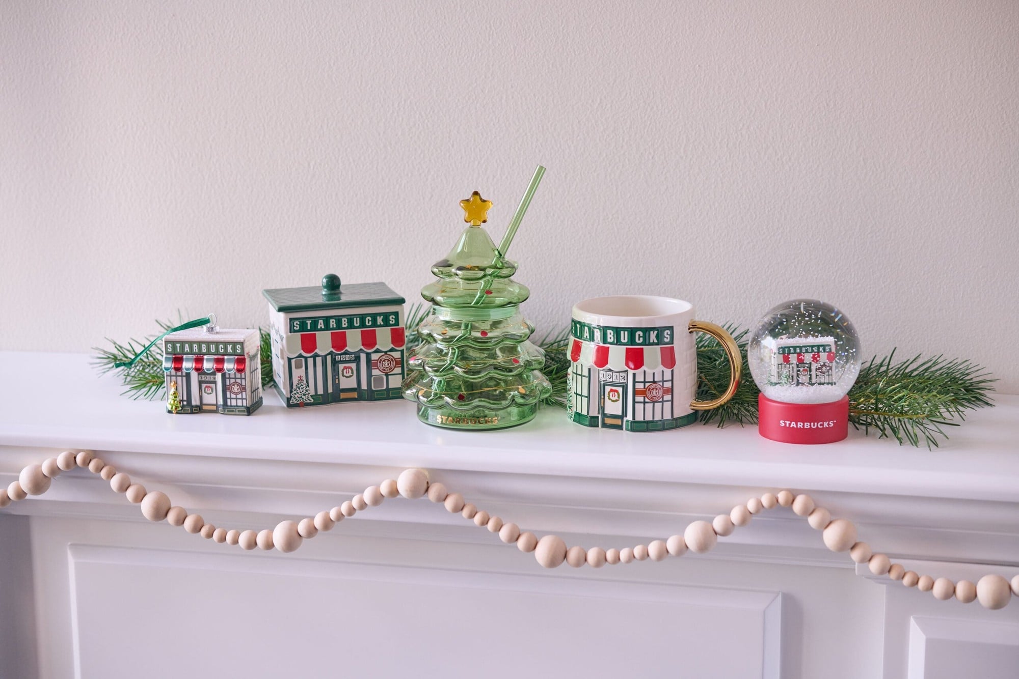 Holiday Village pieces on a mantel: a ceramic Starbucks coffeehouse, a storefront mug, an ornament, a glass tree cup and a snow globe