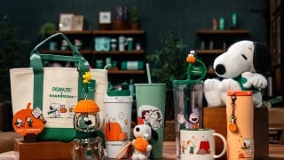 The Starbucks and Peanuts fall 2026 collection: Snoopy glass cold cup, tumblers, mugs, a plush, tote, and charm on a wood table