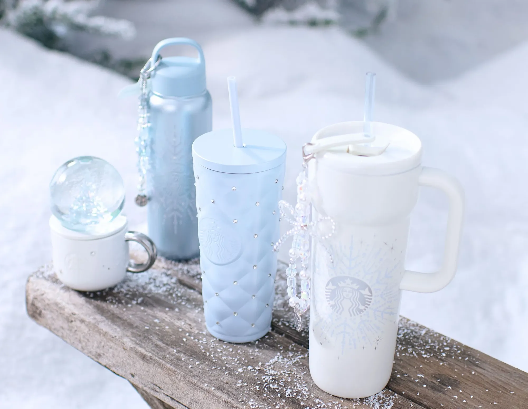 Winter Wonderland drinkware in light blue and white on a snowy bench: a quilted cold cup, water bottles, a mug, a snow globe and a tumbler