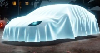A car covered by a glowing white sheet with one eye hole showing sits in a foggy graveyard at night