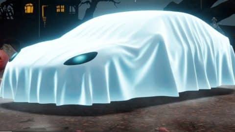 A car covered by a glowing white sheet with one eye hole showing sits in a foggy graveyard at night