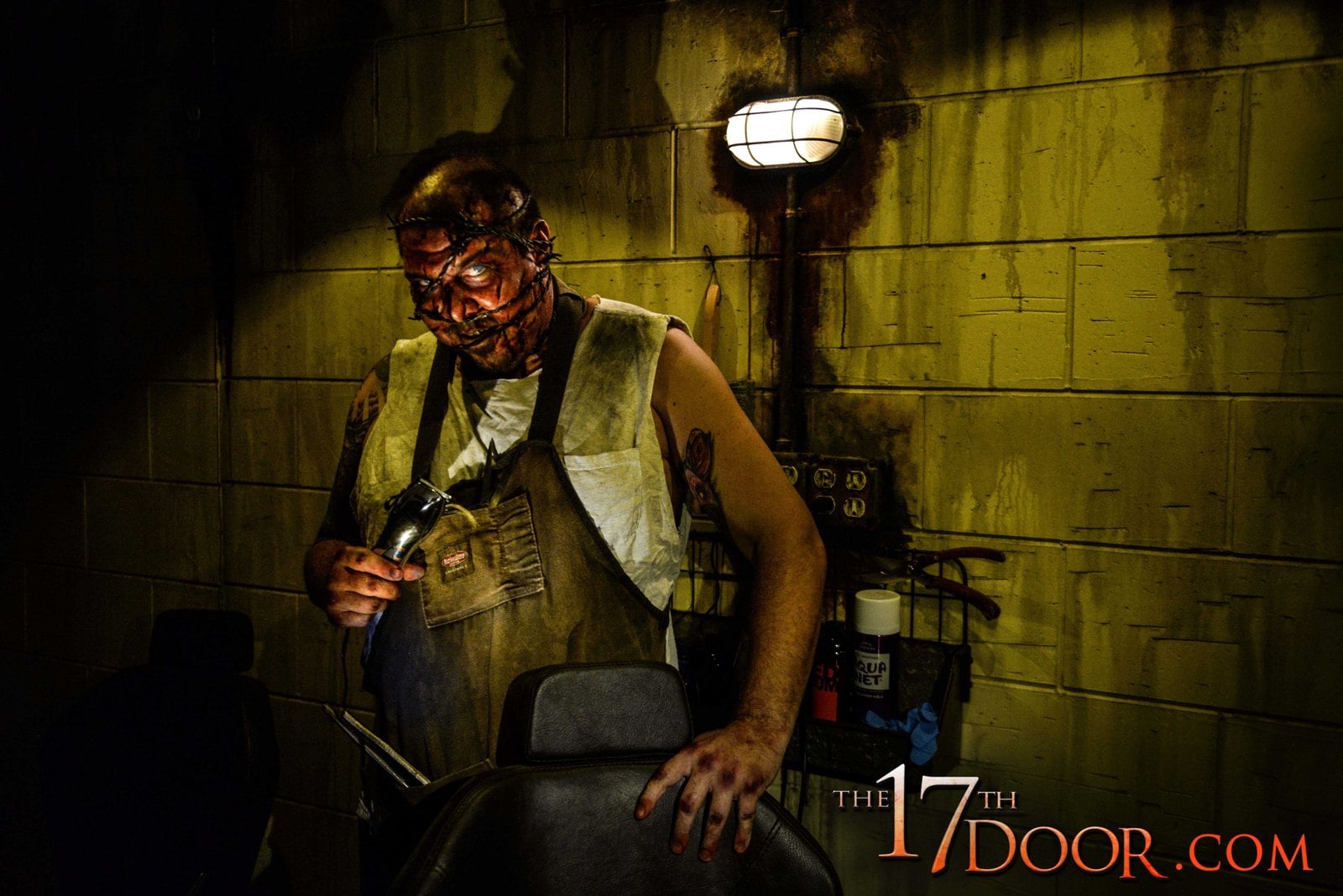 A character in a stained apron with wire wrapped around his head holds hair clippers behind a barber chair in a yellow-lit cinder-block room