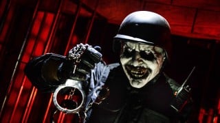 A haunted house character in a black helmet and tactical vest with a painted ghoul face holds up a pair of handcuffs in front of red-lit bars