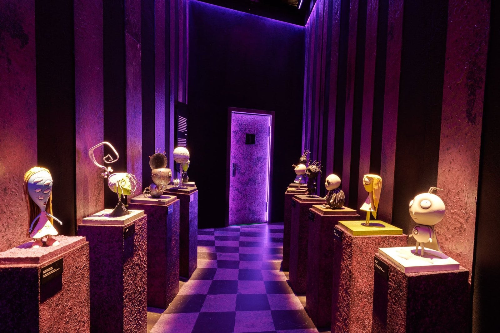Busts of characters on pedestals along both sides of a room with a purple checkered floor and a door at the far end