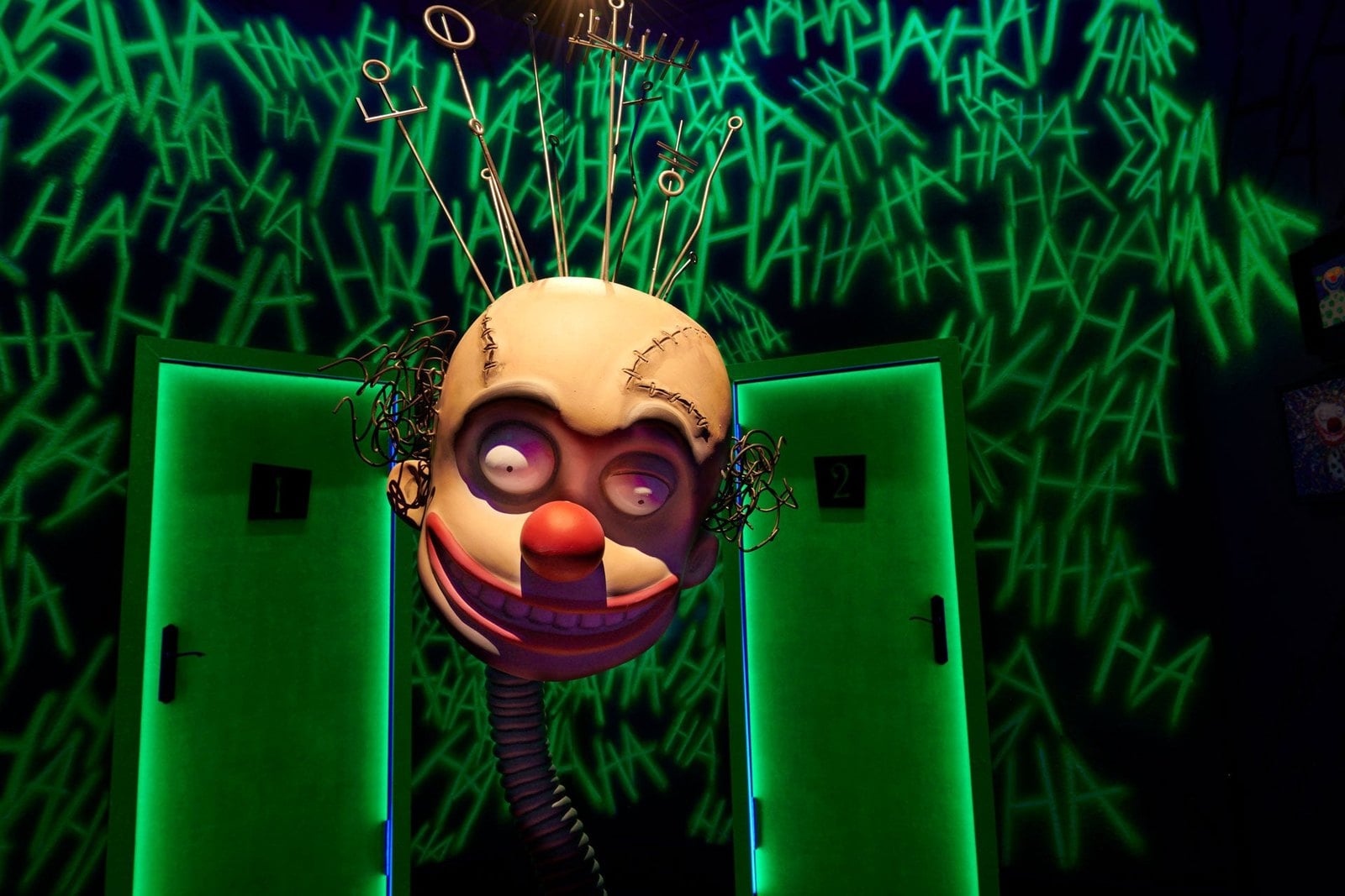 A sculpture of a clown-like head with wires sticking out of its scalp, between two glowing green doors
