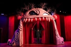 An archway shaped like a pale pink monster's face with one large eye and a row of teeth, with a man in black standing between red curtains