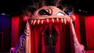 An archway shaped like a pale pink monster's face with one large eye and a row of teeth, with a man in black standing between red curtains
