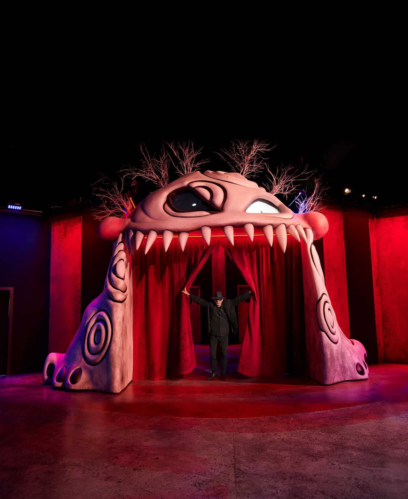 An entrance arch shaped like a pale pink monster's face with one large eye and a row of teeth, with a man in black standing between red curtains
