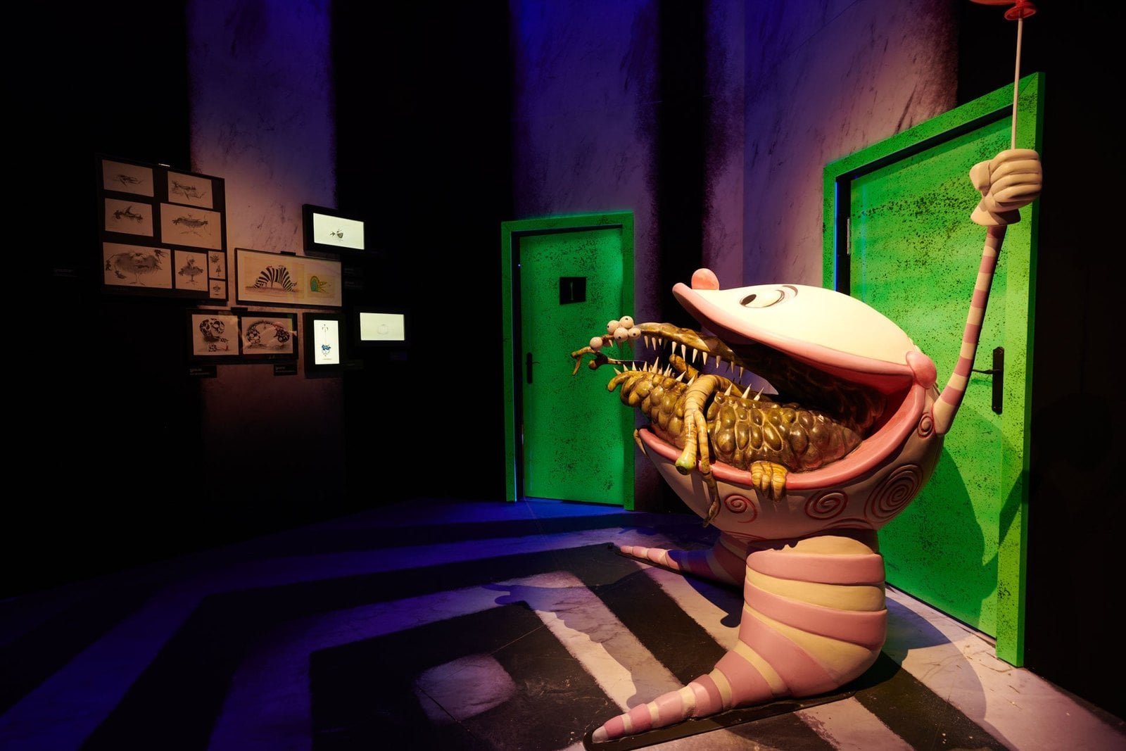 A pink striped creature with an open mouth holding a spiky creature, beside two green doors and a wall of framed drawings