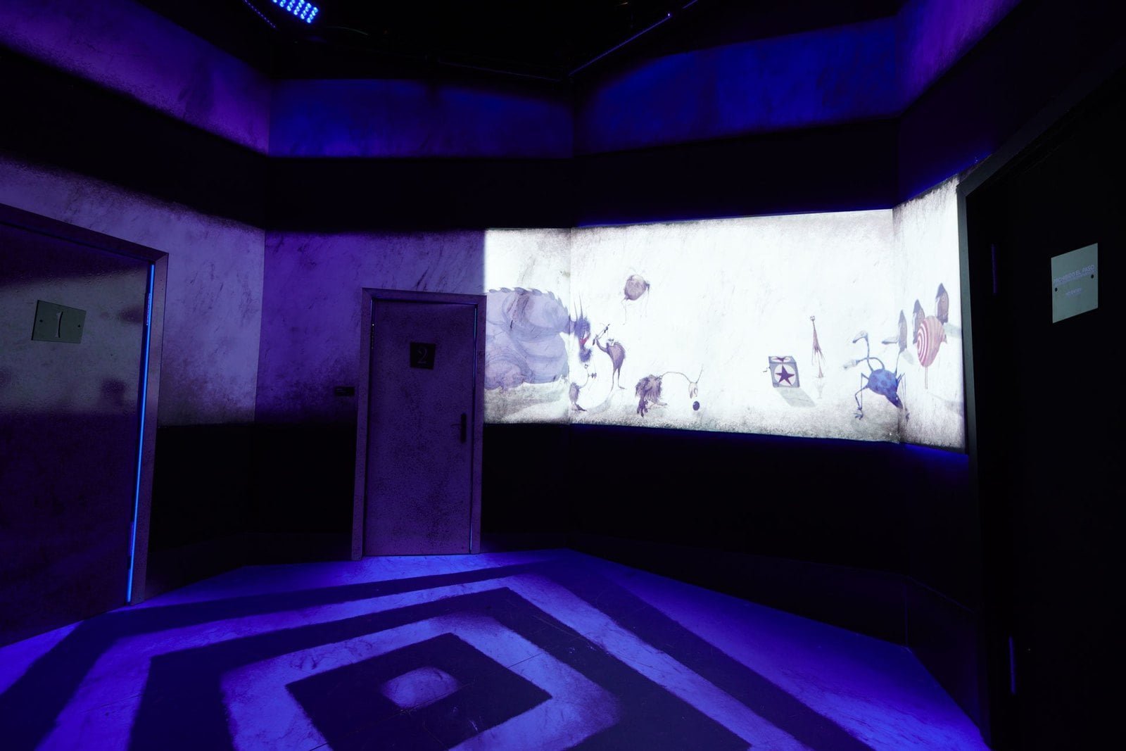 A dark room with a wide screen showing an animated drawing and a door at the left