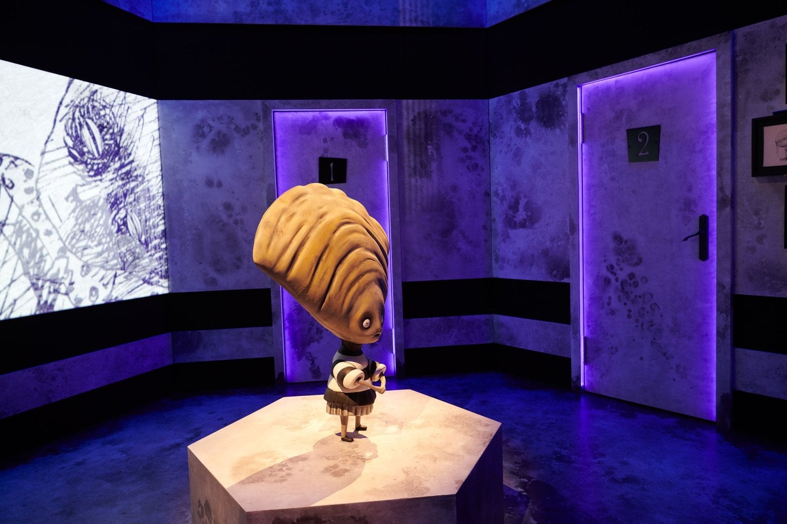 A small figure with a large shell-shaped head stands on a pedestal in a room with drawings projected on the walls