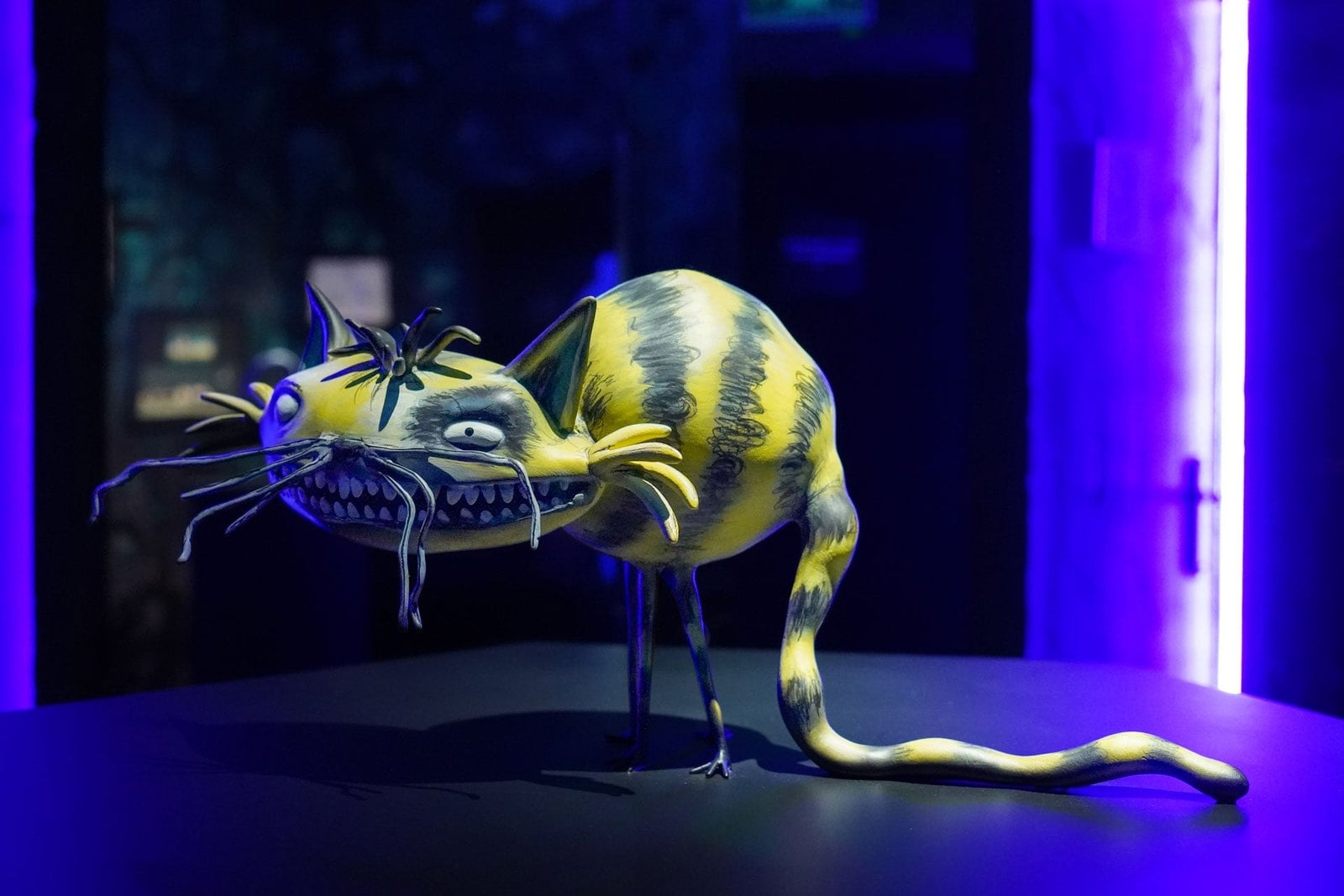 A sculpture of a yellow-and-blue striped cat with long whiskers and a long tail under blue light