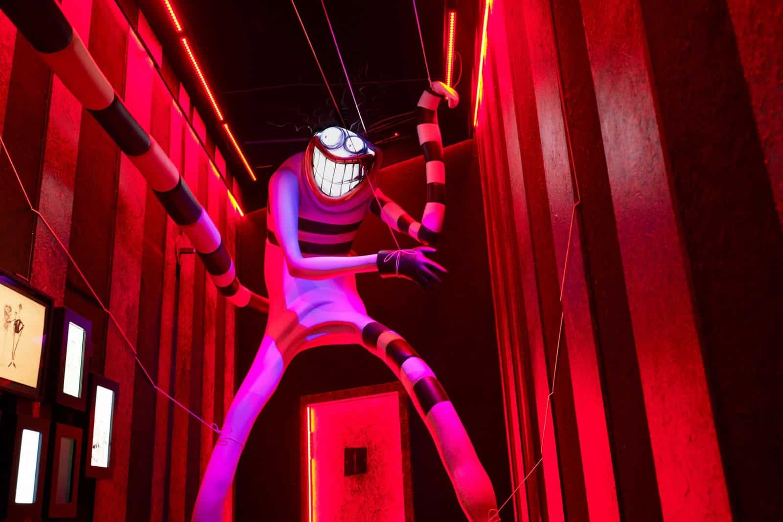 A tall figure with a wide toothy grin and long striped arms stands over a red doorway in a red-lit room