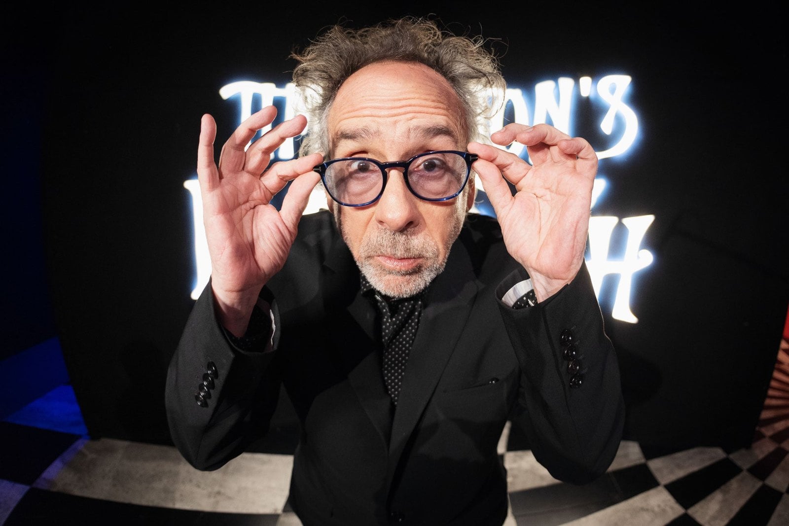Tim Burton holds his glasses with both hands in front of a lit sign