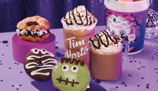 A filled croissant, a mummy donut, a green Frankenstein donut, two chocolate drinks with whipped topping, and a purple bucket printed with a moonlit graveyard, on a purple table