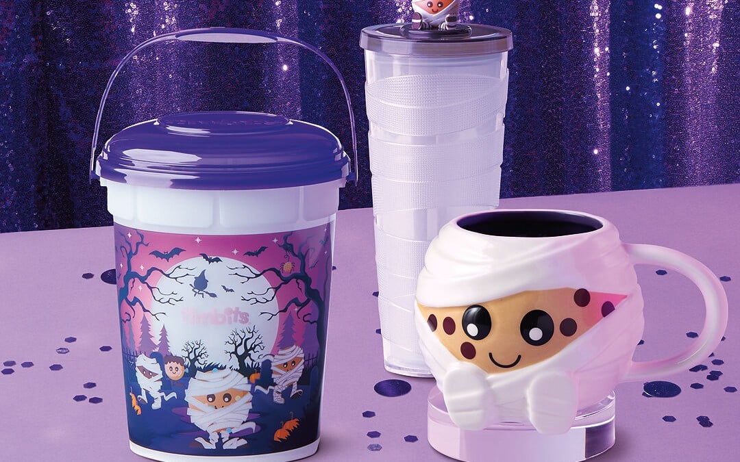 A plastic bucket with a purple lid and a moonlit graveyard scene of small mummy characters, beside a clear tumbler and a mummy-shaped mug