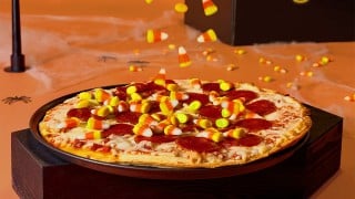 A pepperoni pizza topped with candy corn on a dark wood block, with more candy corn falling onto it against an orange background
