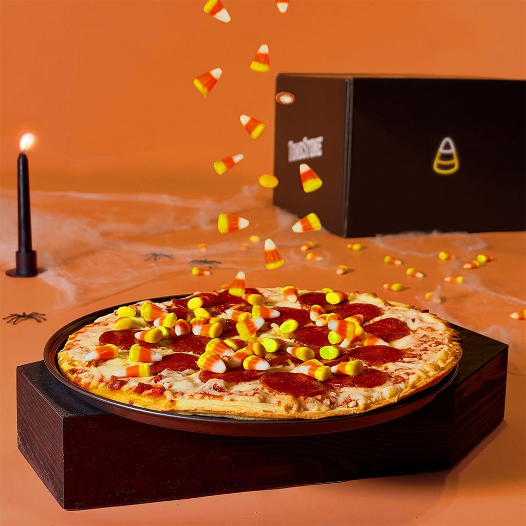 A pepperoni pizza topped with candy corn in front of a black Tombstone box with a candy corn outline, beside a black candle and cobwebs
