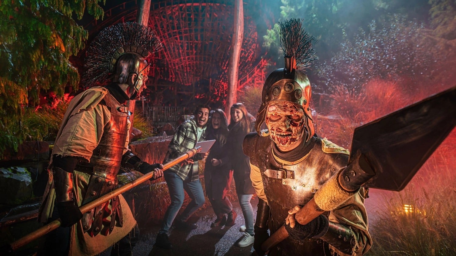 Two mutated Trojan soldiers with spears confront laughing guests in the red-lit DesTroy scare zone at Toverland Halloween Nights, with a roller coaster behind them