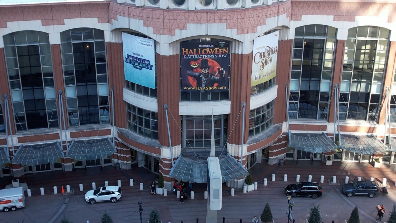 Three large trade show banners hanging above the glass entrance of a brick convention center, seen from above