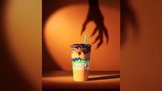 An orange smoothie in a clear cup, topped with dark purple boba and a green straw, in front of an orange wall with the shadow of a clawed hand