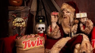 A zombie Santa Claus in a fur-trimmed red suit holds a TWIX bar in one bandaged hand and a stack of cash in the other, beside a burlap sack of TWIX bars and a skull nutcracker