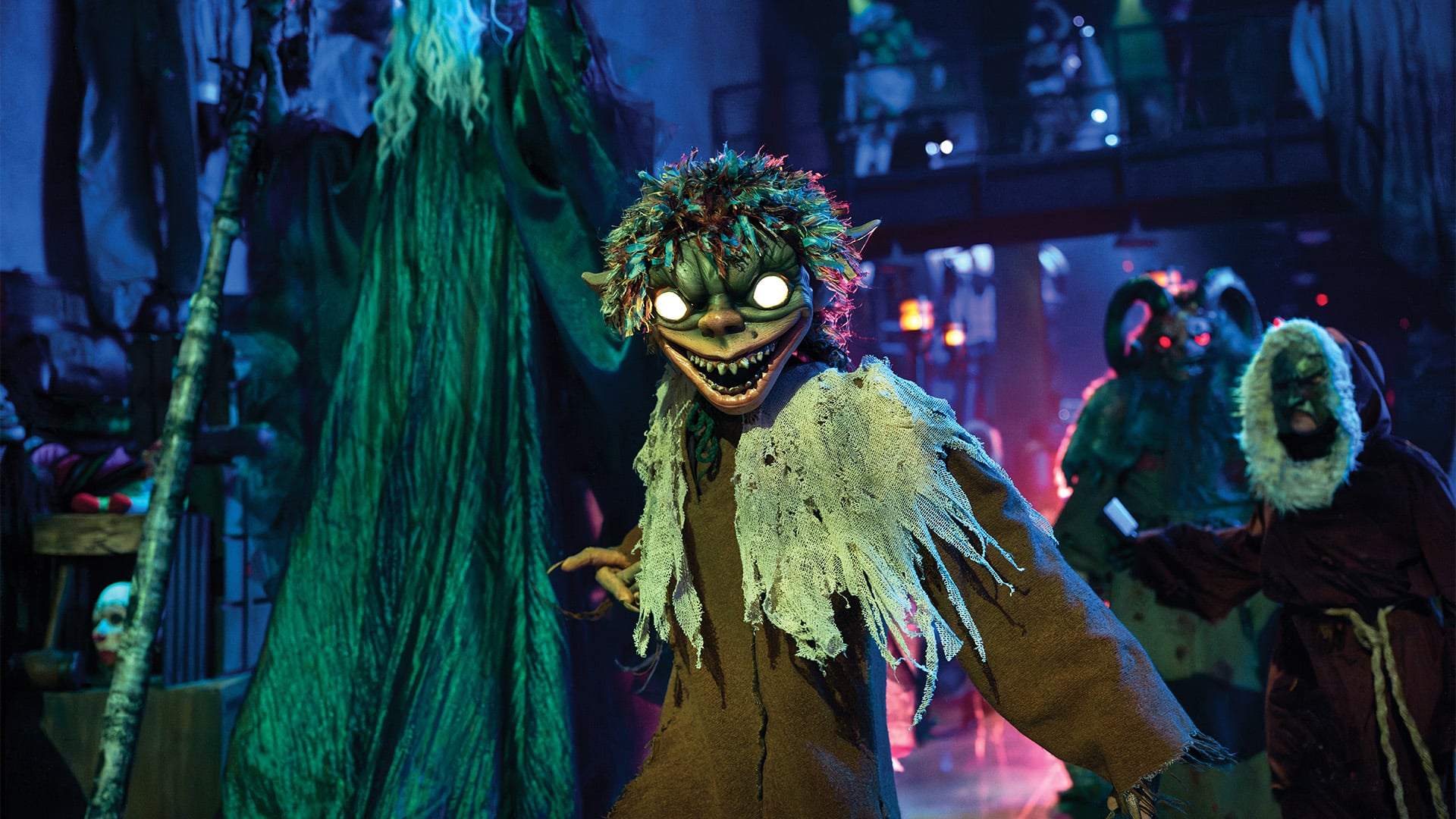 A grinning goblin creature with glowing white eyes and a ragged burlap costume, with horned and hooded creatures behind it in the venue's dim common area