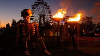 A giant seated skeleton beside two flaming fire pots, with a Ferris wheel against the dusk sky behind it
