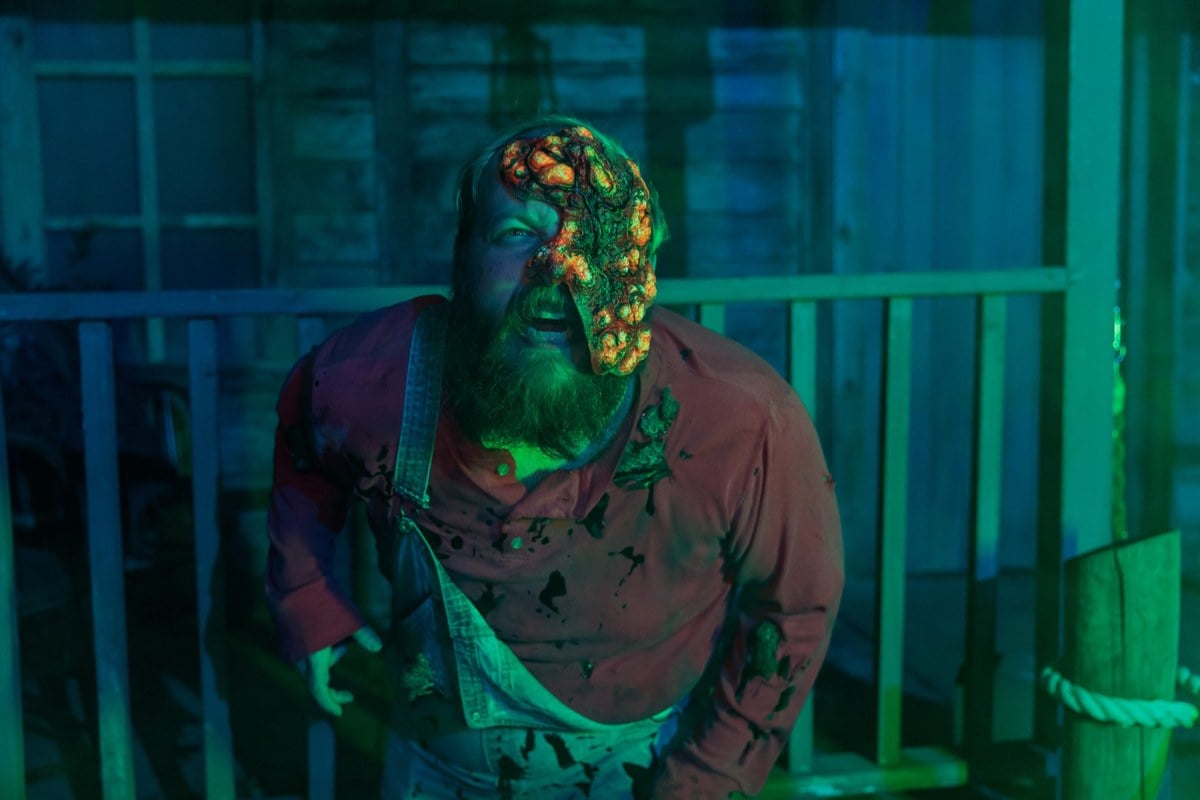 A man with fungus-covered skin and a bloodied apron stands on a green-lit porch