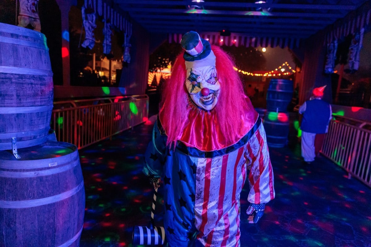 A clown in a striped suit and pink wig walks a lit carnival midway lined with barrels