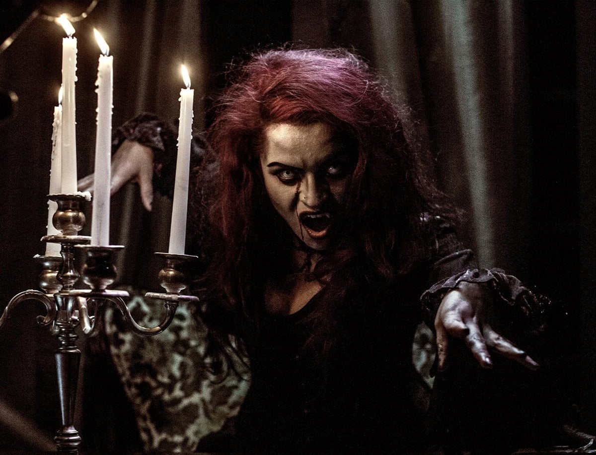 A snarling woman with wild red hair reaches past a lit candelabra in a dark room