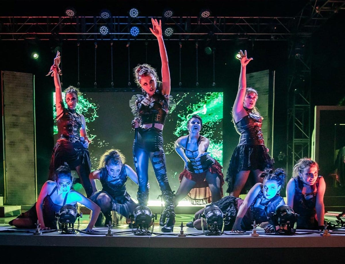 Dancers in dark costumes pose on a stage under green and blue lights