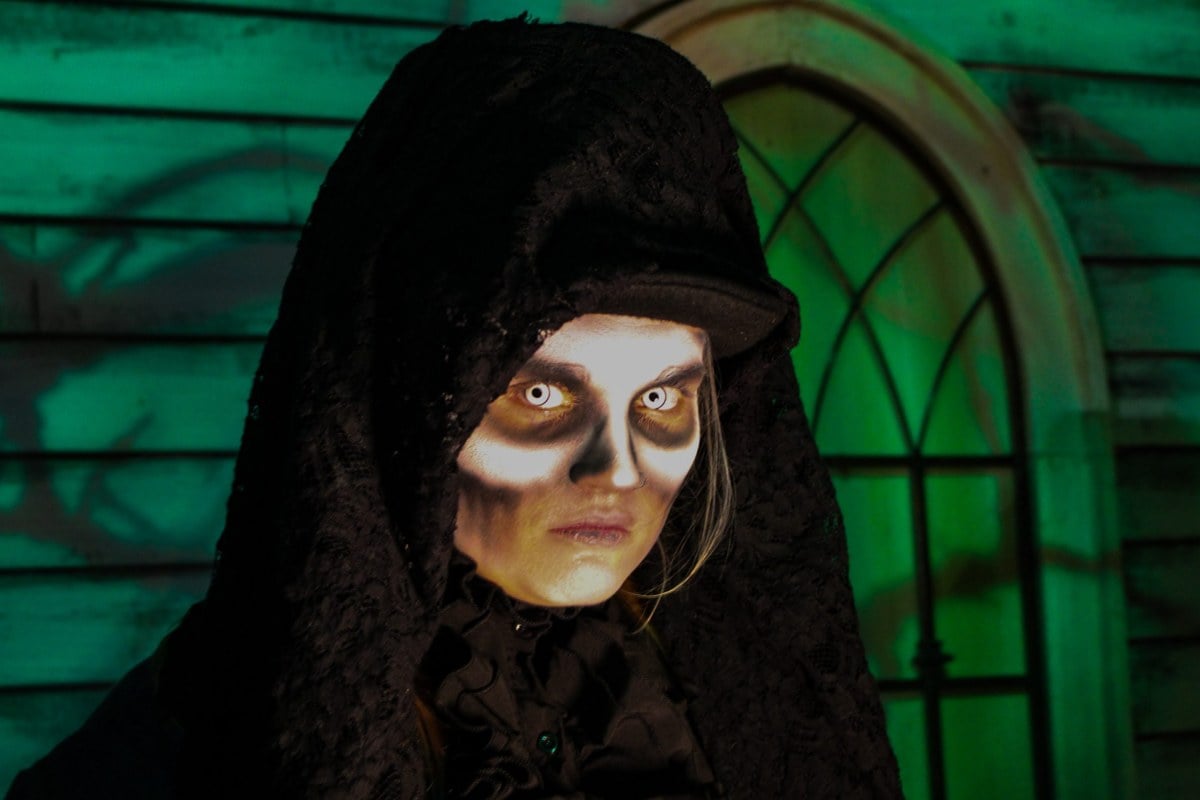 A pale hooded figure with dark-ringed eyes stands in front of an arched green window