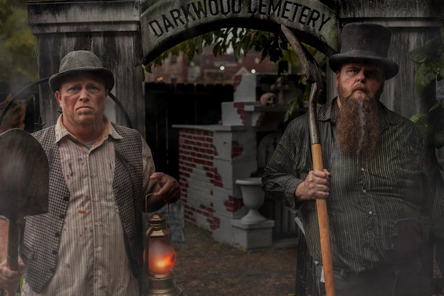 Two men in old-fashioned work clothes hold a shovel, a lantern, and a scythe under an arch that reads DarkWood Cemetery