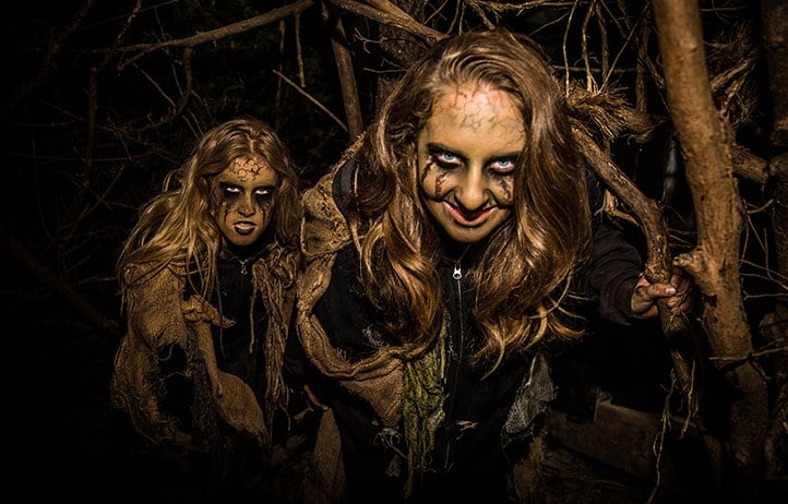 Two performers in cracked face paint and burlap rags crouch among bare branches at night