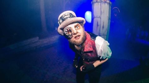 A performer in a white top hat with goggles, a brass eyepiece, and a red vest leans toward the camera in blue fog at night
