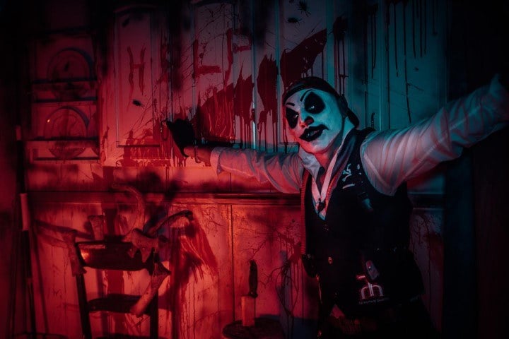 A performer in clown makeup and a black vest spreads both arms in a room lit red