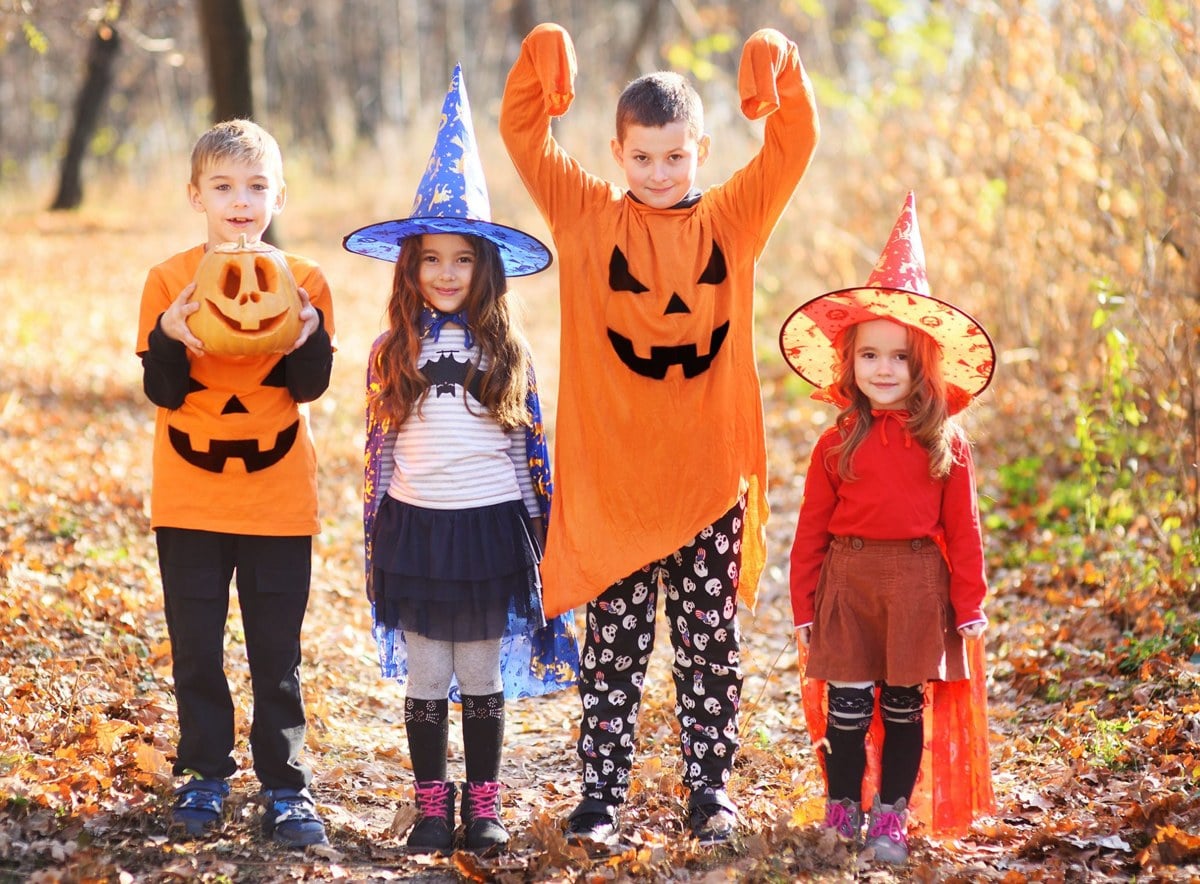 Four children in pumpkin and witch costumes stand in fallen leaves in a sunny forest