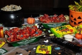 Two trays of chicken wings in a red glaze dotted with candy pieces on a Halloween table with a cauldron of popcorn, a jack-o'-lantern pail and Sour Patch Kids candy