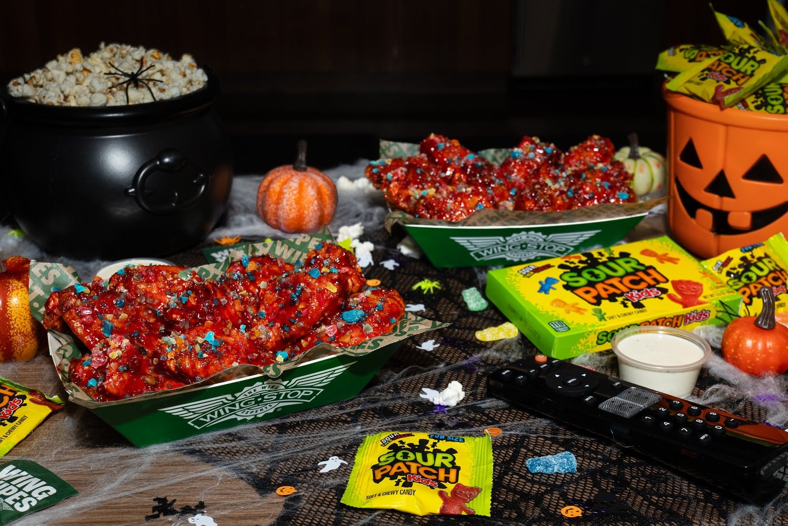 Two trays of chicken wings in a red glaze dotted with candy pieces on a Halloween table with a cauldron of popcorn and a jack-o'-lantern pail
