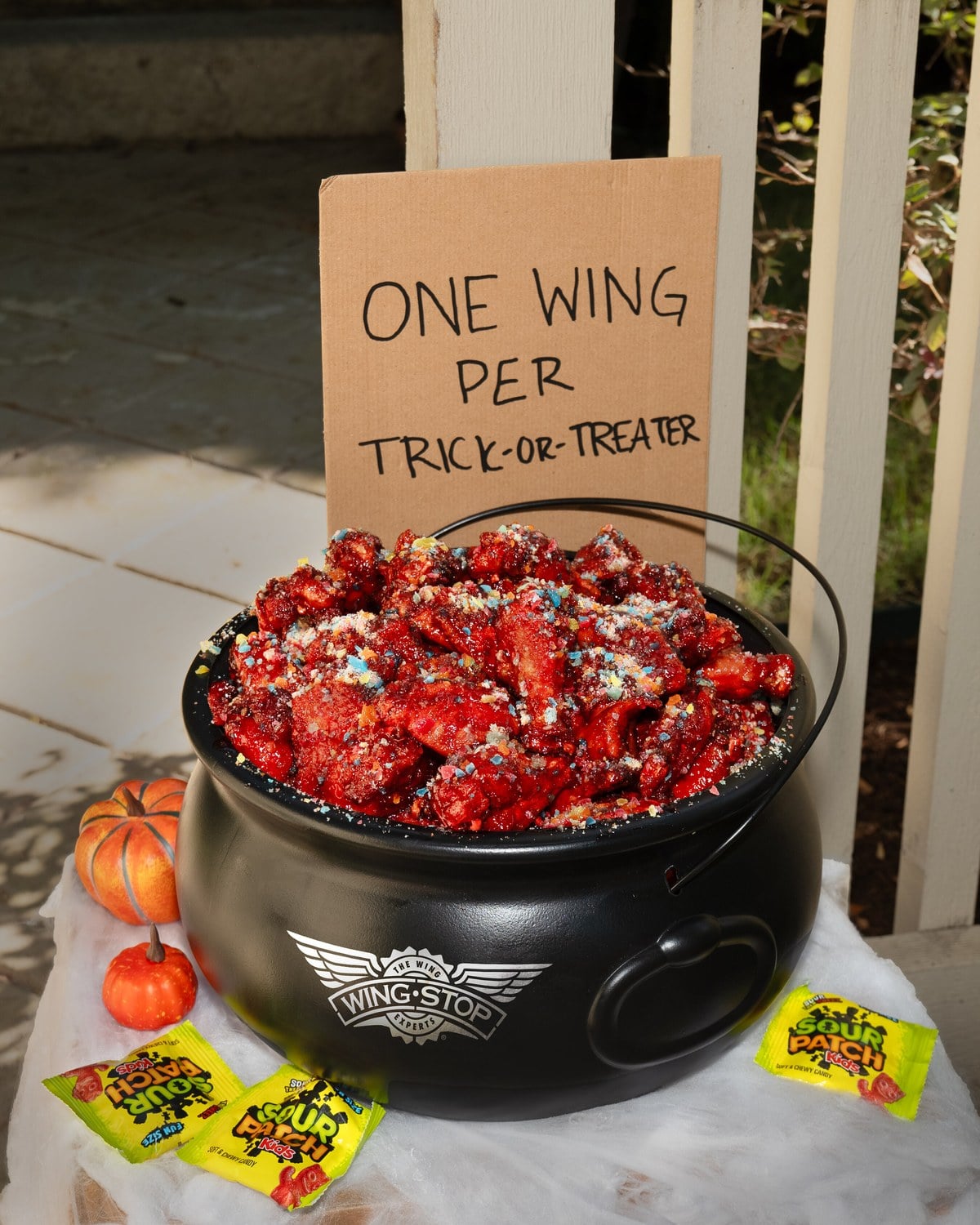 A black cauldron full of red-glazed chicken wings sits on a porch step under a cardboard sign that reads One Wing Per Trick-or-Treater