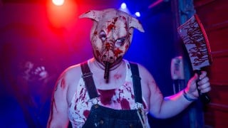 A person in a bloody pig mask, overalls, and a stained tank top holds up a bloody cleaver under red and blue lights