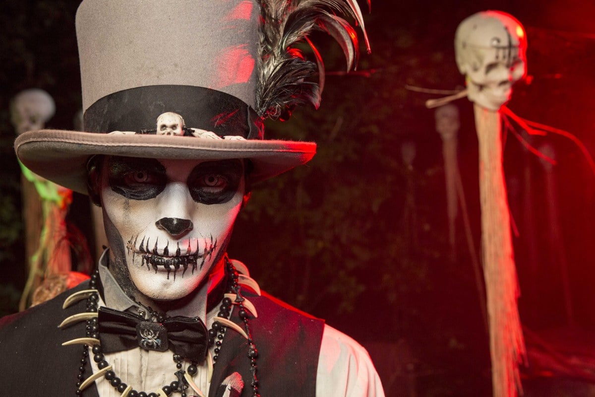 A performer in a top hat with a skull-painted face and a necklace of teeth, with a skull on a pole behind him